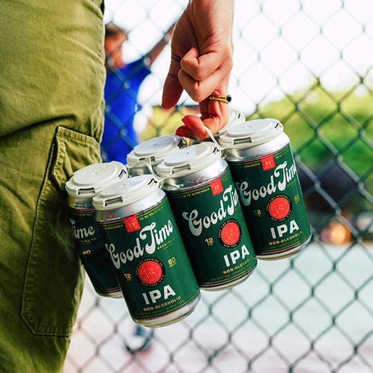 Someone holding a 6-Pack of Good Time Brewing's NA IPA beside them while looking through a fence.