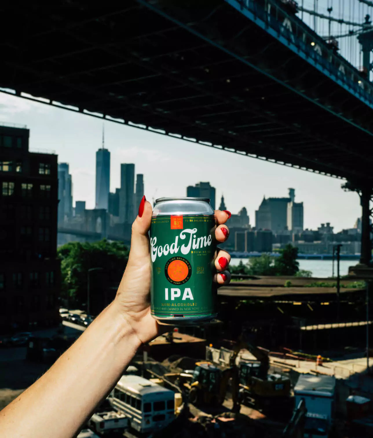 A woman's hand holding a Good Time Brewing IPA Non Alcoholic Beer in front of the New York City Skyline and a New York City bridge.