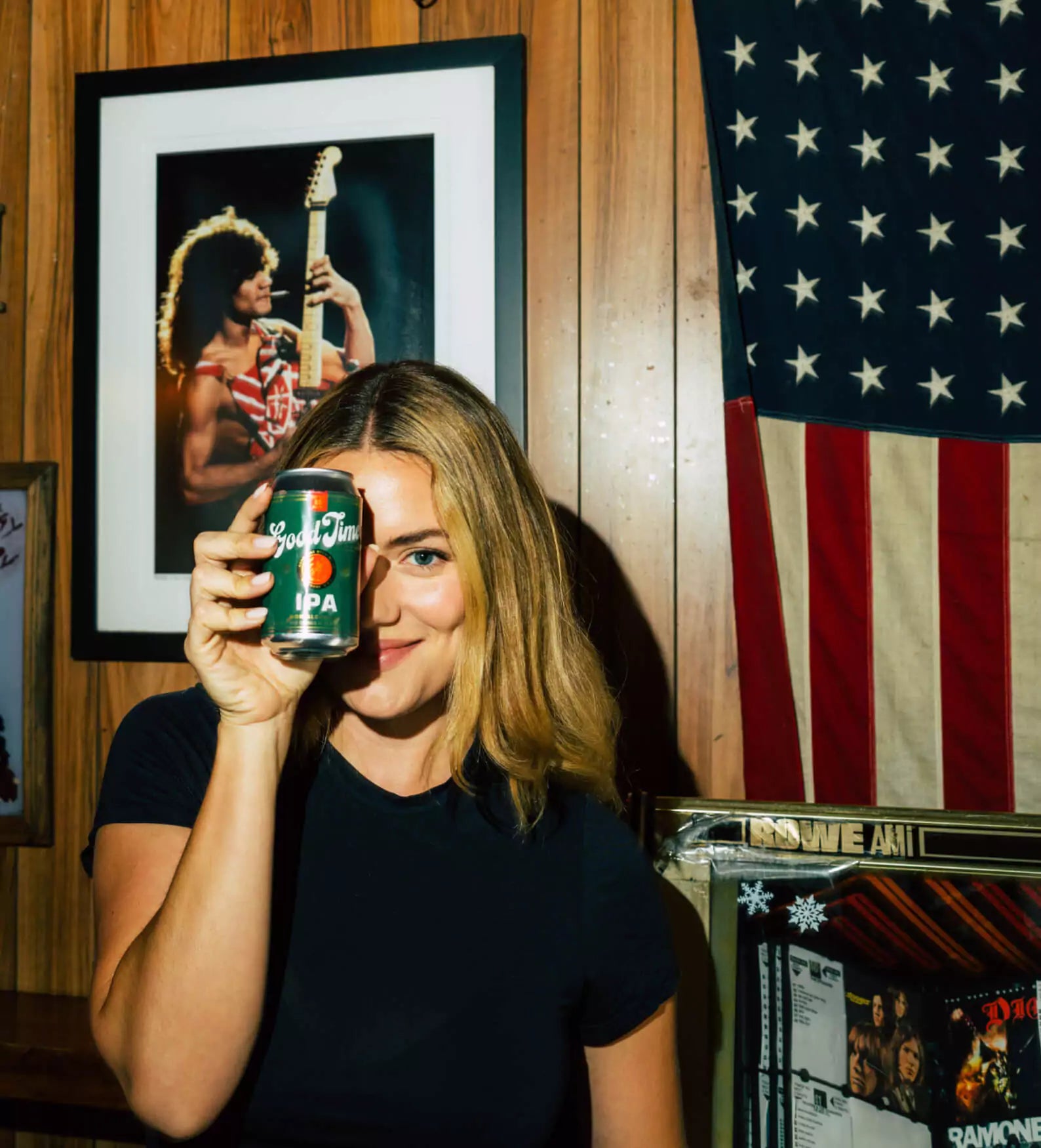 A woman holding a 'Good Time IPA Non Alcoholic Beer' in front of her frace. Behind her is a guy playing guitar, as well as an american flag, and to the bottom right of the photo is what appears to be an old school juke box.